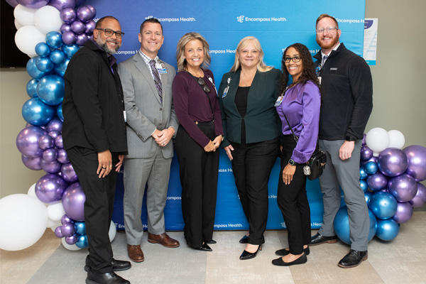 Encompass Health Toledo employees stand smiling in front of a blue 