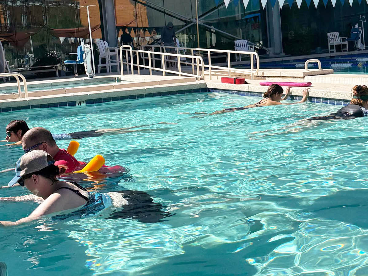 Adults in a pool practice floating, with colorful kickboards nearby. Bright, clear water reflects a sunny atmosphere, creating a harmonious scene.