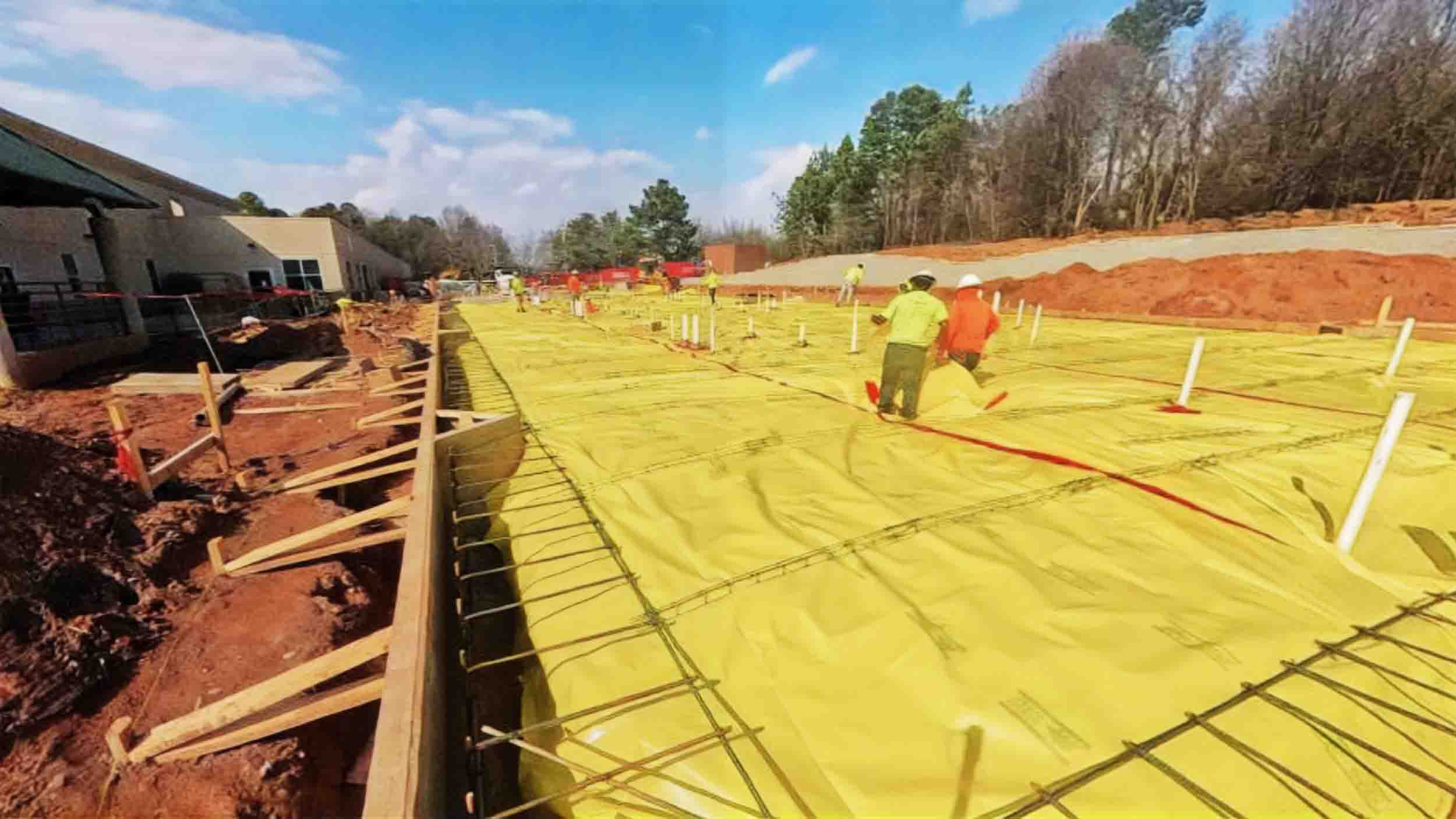 Construction crews work on the AnMed building expansion, installing vapor barriers and rebar across a large foundation area surrounded by soil and equipment.