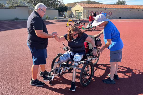 A female amputee on a recumbent tricycle is assisted by two people on a sunny outdoor track. The atmosphere is supportive and encouraging.