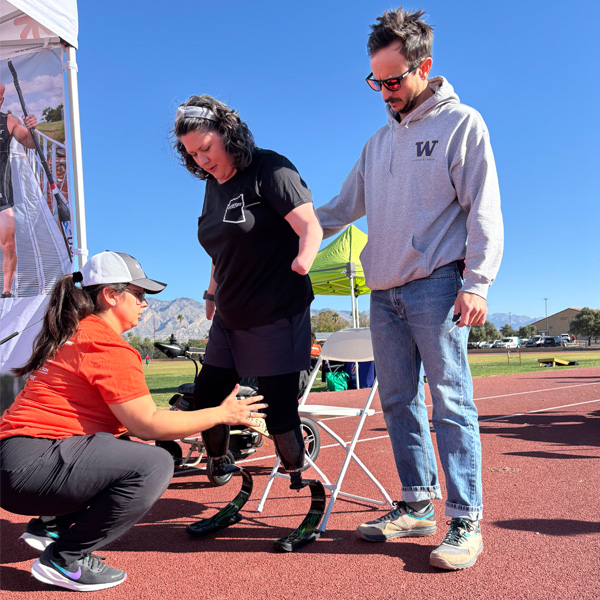 A woman with prosthetic legs is assisted by a man and a woman on a track. The setting is sunny with mountains in the background, conveying perseverance.