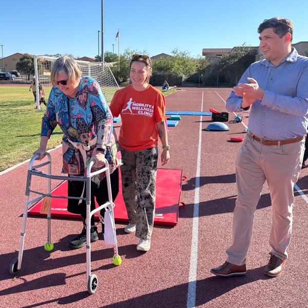 A smiling woman in a colorful shirt uses a walker on a track, assisted by a woman in a red shirt. A man in a blue shirt claps supportively nearby.