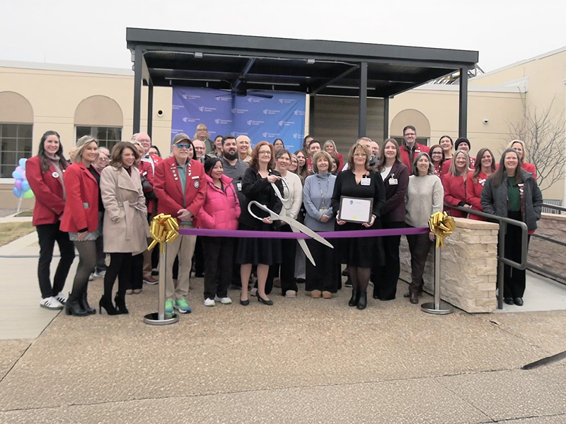 A group of people is gathered for a ribbon-cutting ceremony outside a building. They stand behind a large purple ribbon with oversized scissors and a gold bow, smiling and celebrating.