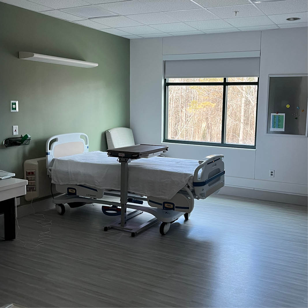 A clean, sunlit hospital room with a single bed, white sheets, and a bedside table. A large window shows trees outside, creating a calm, serene atmosphere.