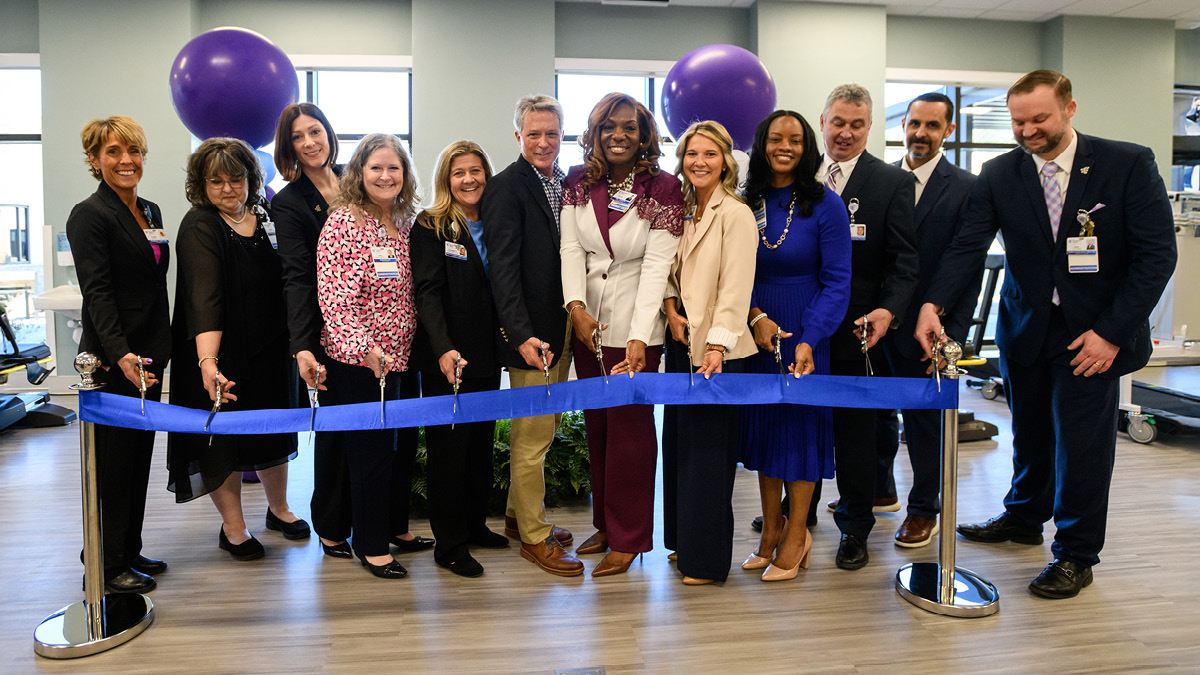 A group of diverse professionals smiling while cutting a blue ribbon at an opening ceremony, two large purple balloons in the background.