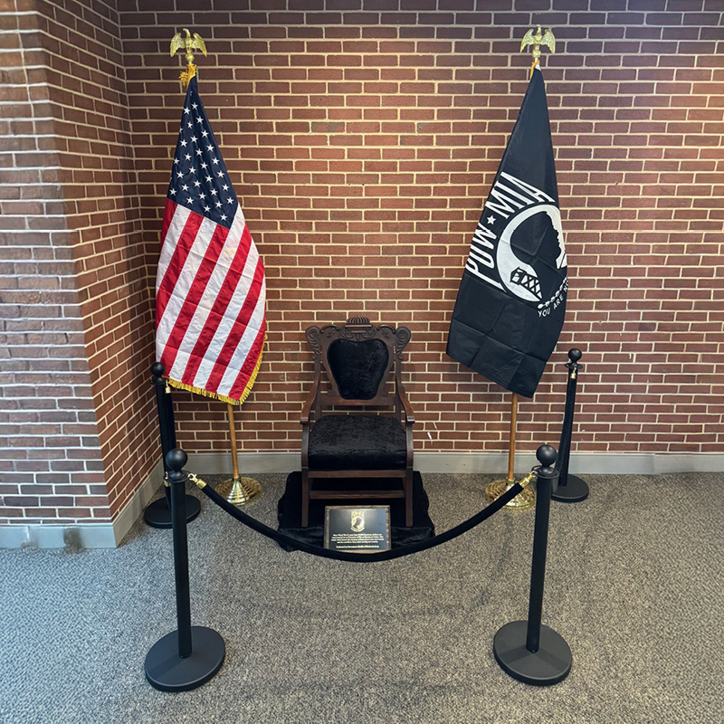 A wooden chair with a black velvet cushion is displayed between the U.S. and POW-MIA flags. Velvet ropes and a plaque enhance the somber setup.