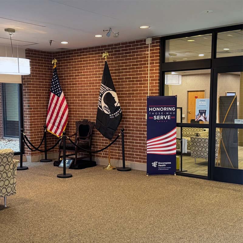 A room with brick walls features the U.S. and POW/MIA flags beside an empty black chair. A banner reads 'Honoring Those Who Serve,'' conveying respect.