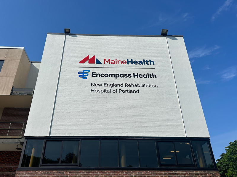 A white building facade displays logos for MaineHealth and Encompass Health, with blue sky above. The sign reads New England Rehabilitation Hospital of Portland.