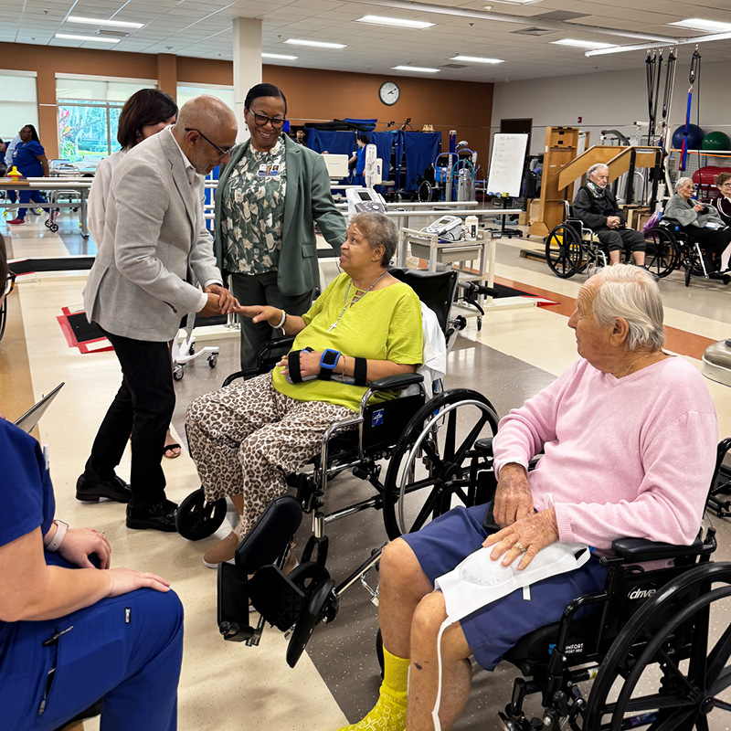 A person in a white blazer warmly shakes hands with a woman in a wheelchair, wearing a bright green shirt. Another man in a pink shirt looks on in a lively rehabilitation center, conveying a sense of community and care.