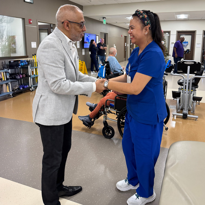 A man in a gray blazer shakes hands with a smiling woman in blue scrubs in a clinic. Wheelchairs and exercise equipment are visible, suggesting a healthcare setting.