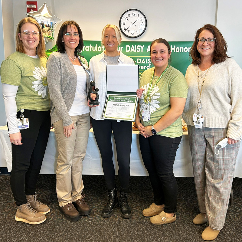 Five women stand smiling in celebration. Four wear green daisy-themed shirts, one holds a DAISY award, and a certificate is visible. Balloons and a congratulatory banner adorn the background.