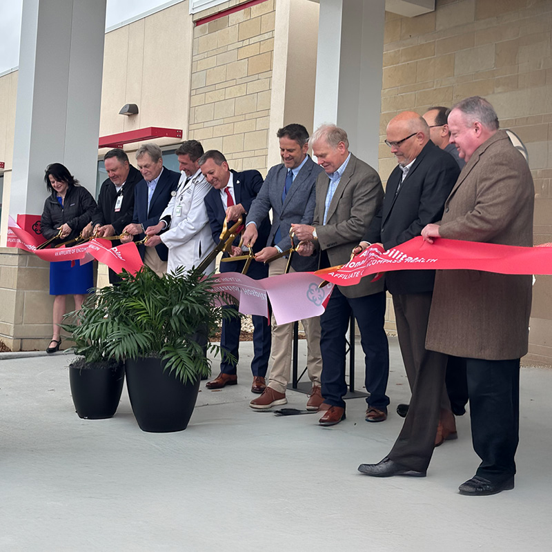 A group of people stand outside a building, smiling as they cut a large red ribbon with oversized scissors. There are potted plants in the foreground.