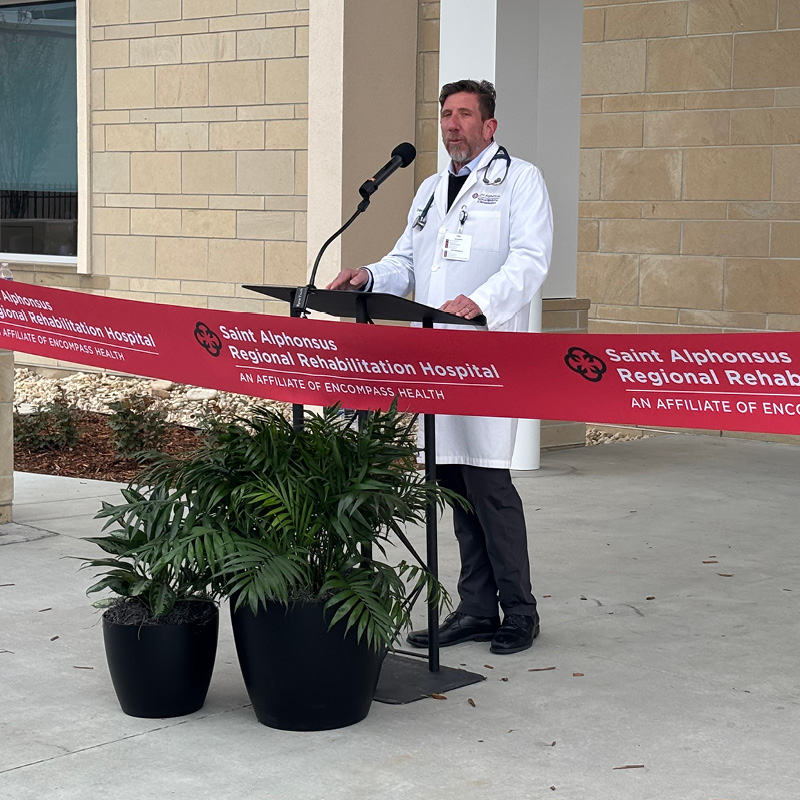 A doctor in a white coat stands at a podium speaking into a microphone during a ribbon-cutting ceremony for Saint Alphonsus Regional Rehabilitation Hospital.