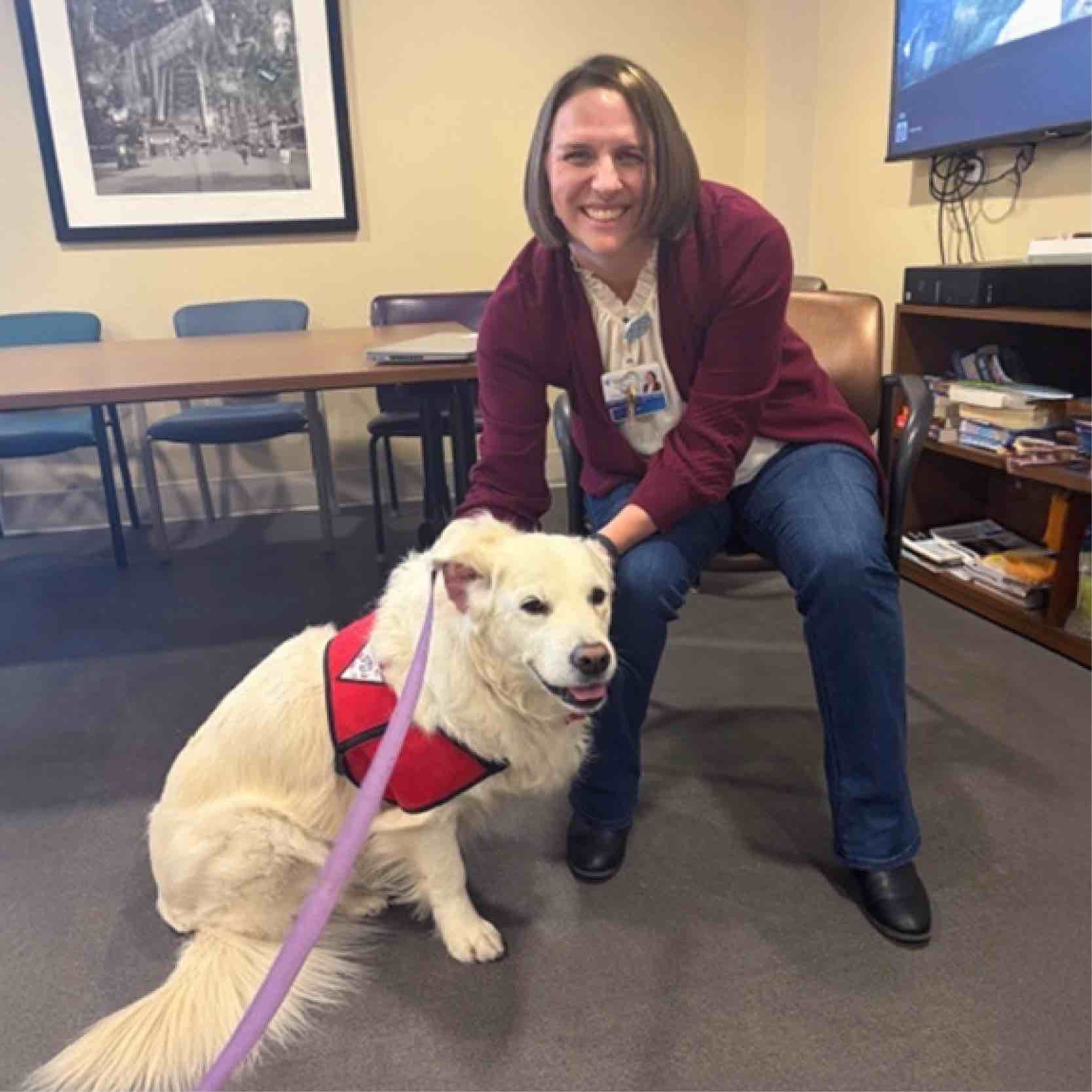 A smiling healthcare employee kneels beside a therapy dog at a rehabilitation hospital, showcasing staff wellness, companionship, and the positive impact of animal-assisted therapy programs.