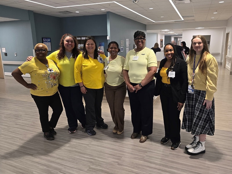 A group of seven people stands smiling in a well-lit hallway, mostly wearing yellow tops, conveying a cheerful and professional atmosphere.