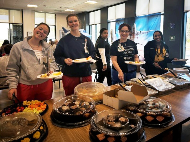 Smiling group of people, holding plates, at a buffet table filled with covered dishes and fruit platters in a bright room with large windows.