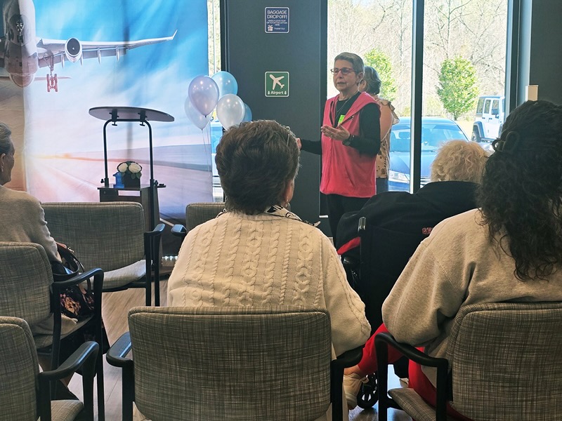 A woman stands speaking in a room with rows of seated people. Behind her is a large airplane mural and balloons.