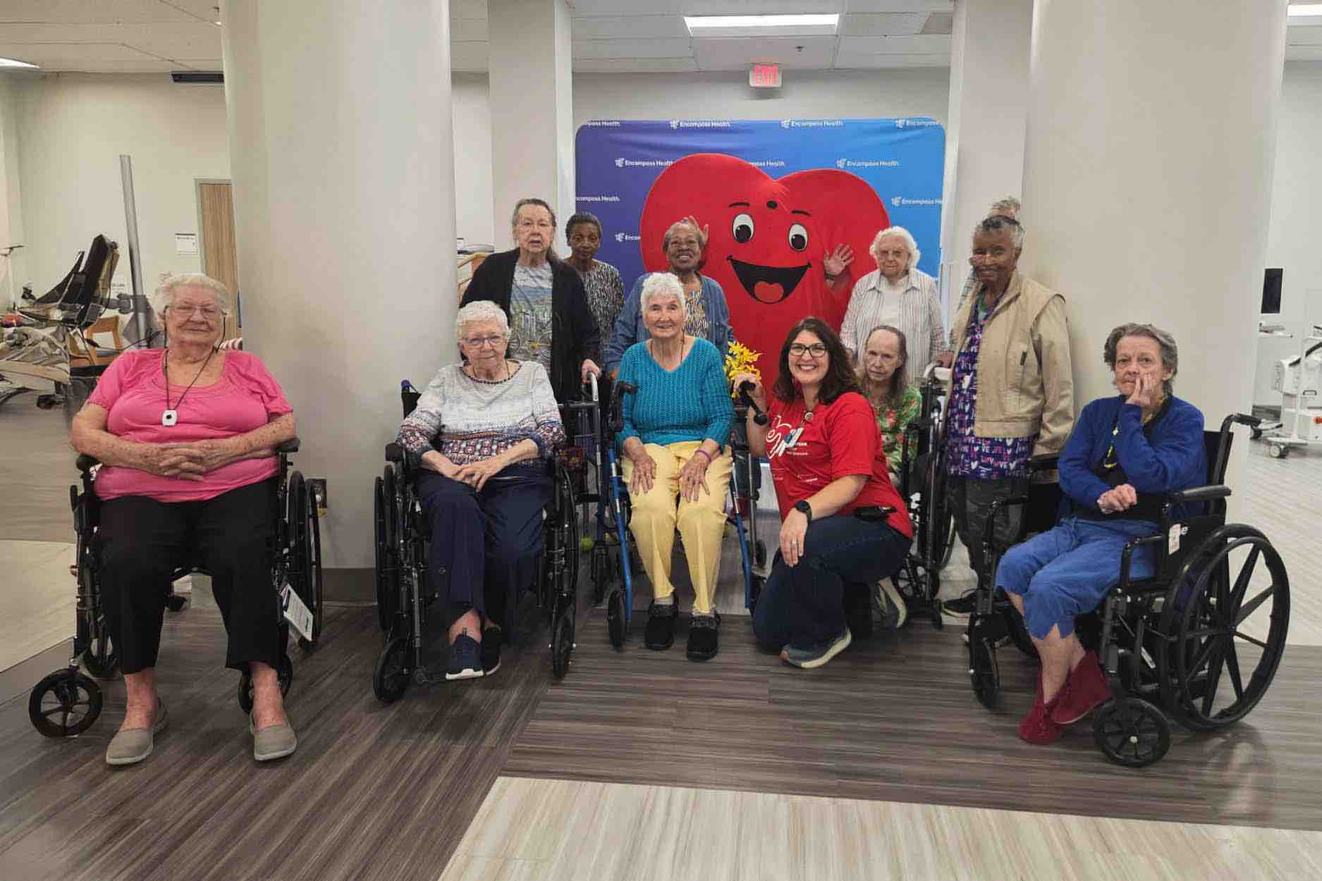Stroke Field Day group photo of rehabilitation patients and staff in a therapy gym, with several wheelchair users and a large red heart mascot promoting stroke recovery and community engagement