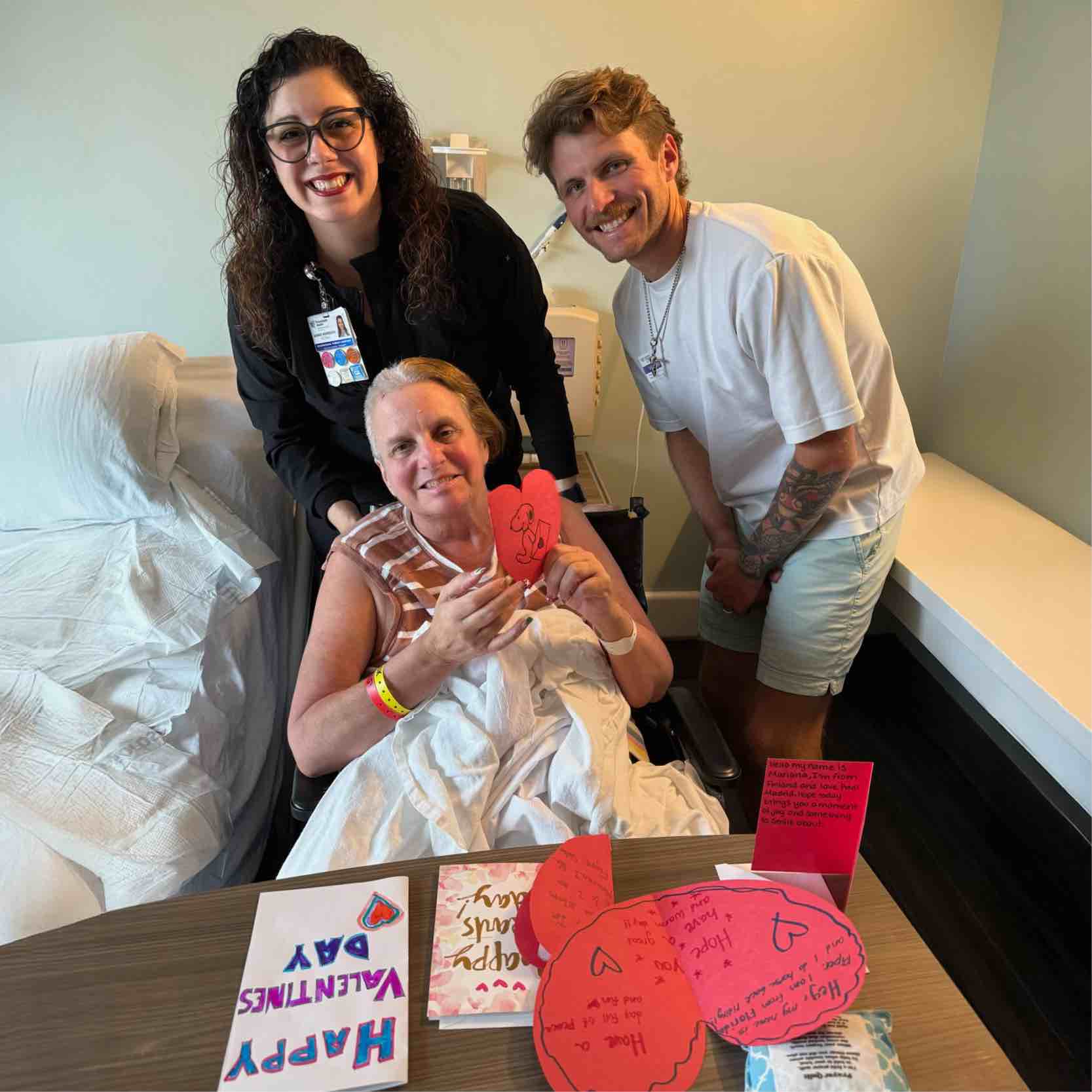A patient sits in a hospital room holding a handmade heart card, with two visitors standing beside the bed. Valentine’s Day cards from Monteverde Academy are displayed on the table.