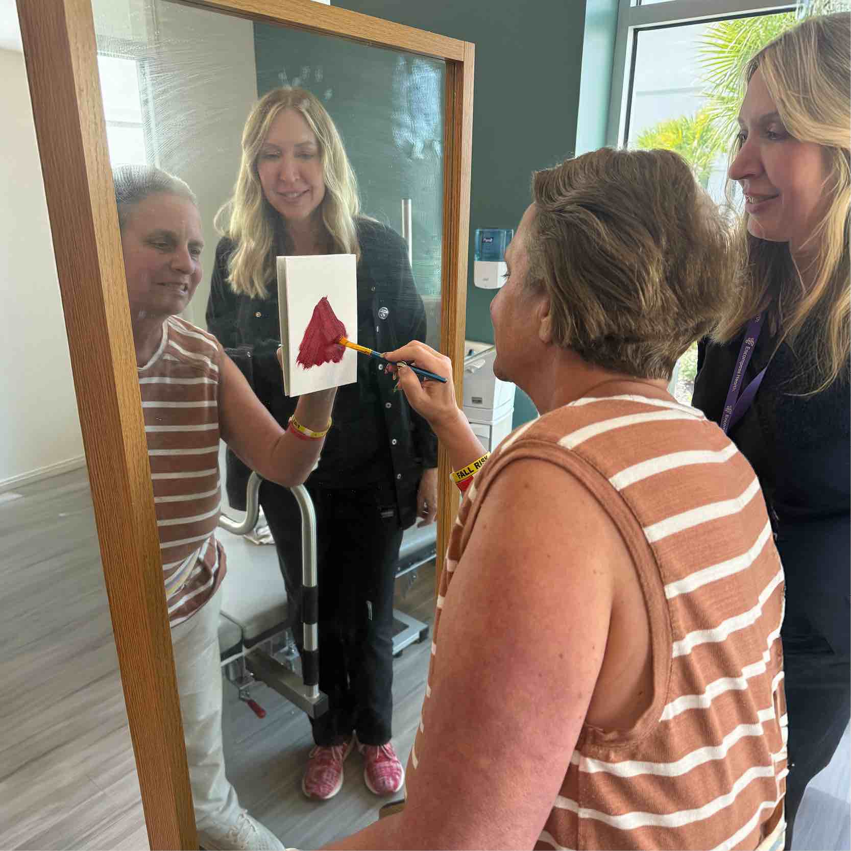 A person paints a red heart on a canvas while seated in front of a mirror, with two individuals standing nearby for support during the Monteverde Academy Valentine activity.