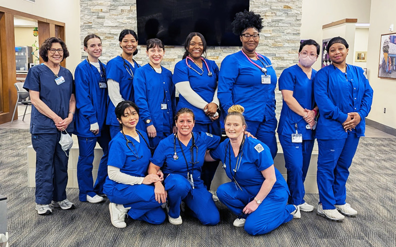 A group of eleven healthcare professionals in blue scrubs pose together in a hospital setting. They appear cheerful and united, exuding teamwork and camaraderie.