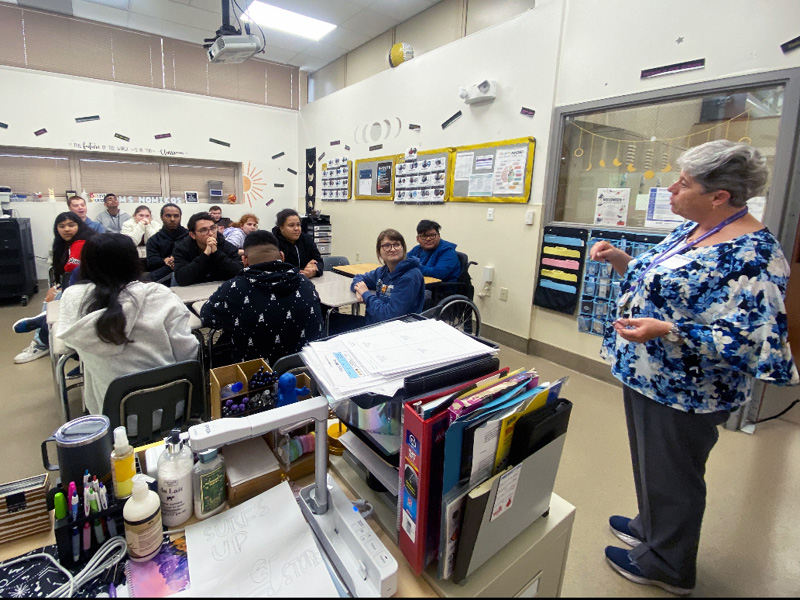 A teacher speaks to a diverse group of students seated at tables in a classroom. The room is bright, with educational posters on the walls and supplies on a desk.