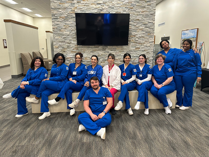 A group of healthcare professionals in blue scrubs sit and kneel together in a well-lit room with a stone wall and a large black screen, smiling warmly.