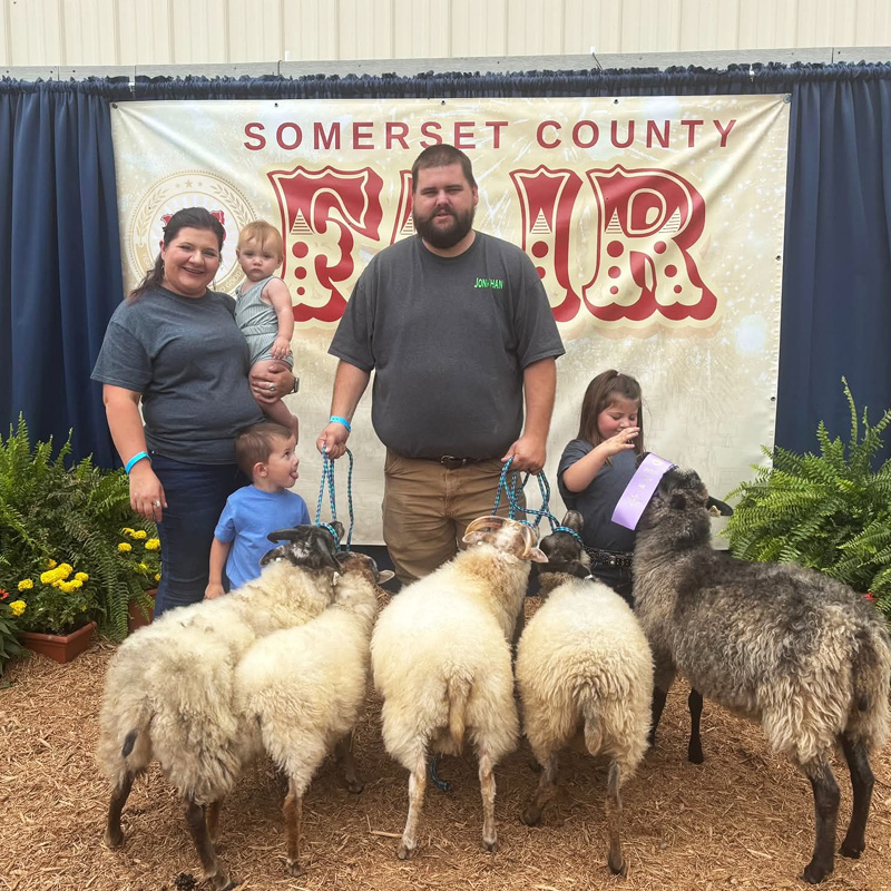Family posing with sheep in front of a 'Somerset County Fair' sign. The group includes adults and children, surrounded by greenery, conveying a joyful rural fair atmosphere.