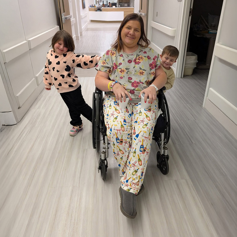 A woman in a wheelchair is joyfully accompanied by two children in a bright hospital hallway. The group is smiling, conveying a warm and happy atmosphere.