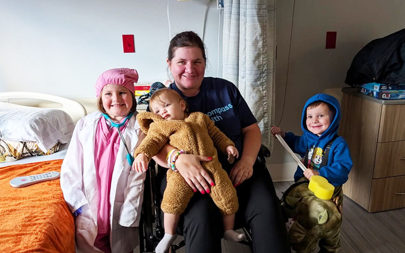 A smiling woman in a wheelchair with three children. One child in a doctor costume, a toddler in her lap in a bear suit, and another child with a toy shovel.