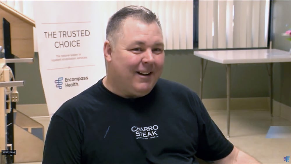 A smiling man in a black "Charro Steak" t-shirt sits in a room with a sign reading "The Trusted Choice" by Encompass Health. The mood is friendly and welcoming.