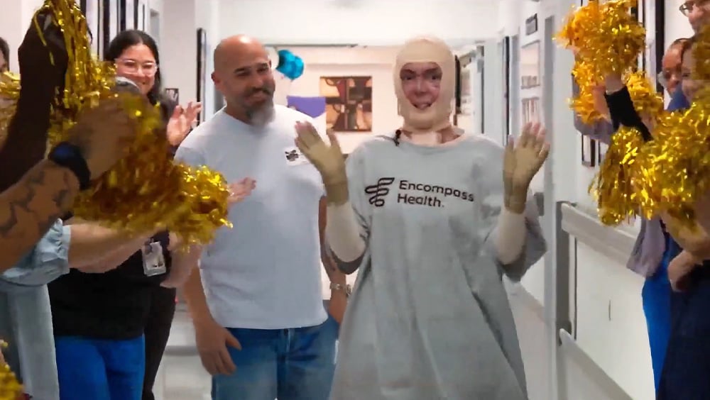 A person in an Encompass Health gown, with facial bandages, walks down a hospital corridor, smiling. Staff cheer with gold pom-poms, conveying a celebratory mood.