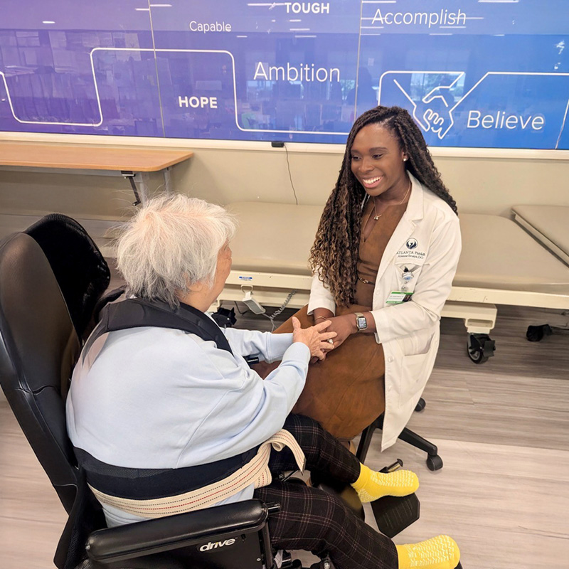 A healthcare professional smiles warmly and holds hands with an elderly patient in a wheelchair. The setting conveys care and empathy.