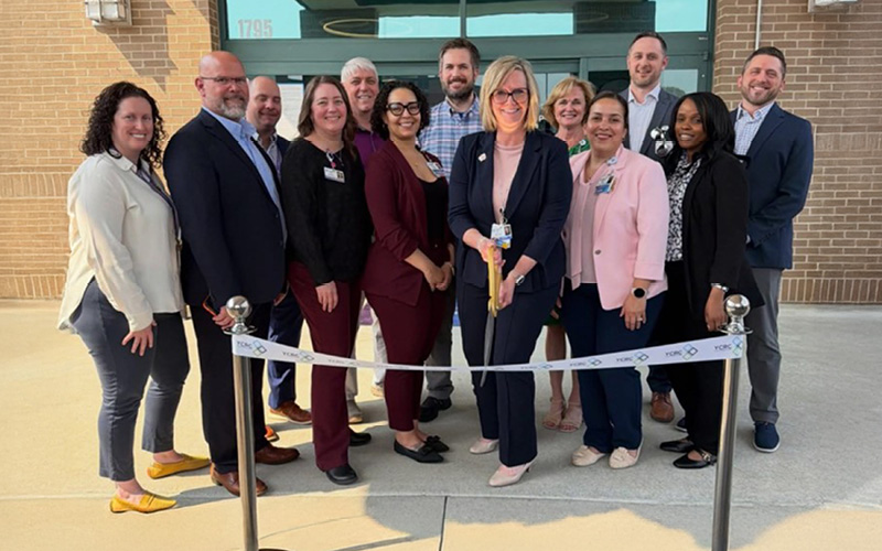 A group of 13 smiling adults stands in front of a building, behind a ribbon marked for a ceremony. The tone is celebratory and professional.