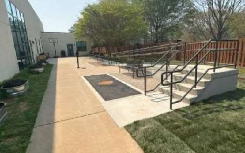 A sunny outdoor pathway features a basketball hoop, ramp with metal railings, and adjacent stairs. Benches and trees create a peaceful setting.