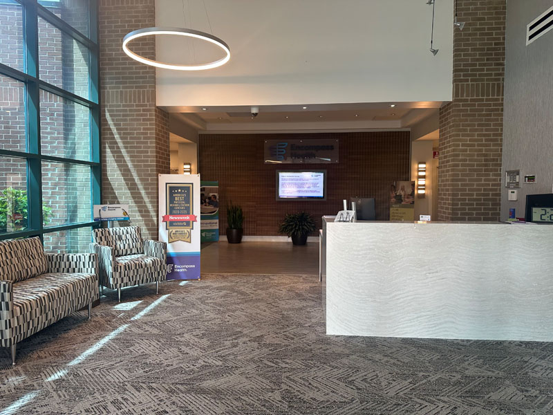 A modern reception area with large windows casting natural light, patterned chairs, a sleek white counter, digital screen, and soothing, neutral tones.
