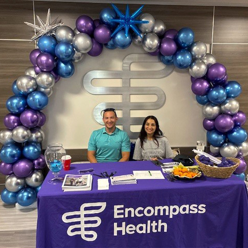 Two people sit at a table with an Encompass Health tablecloth, under a purple, blue, and silver balloon arch. The atmosphere is welcoming.