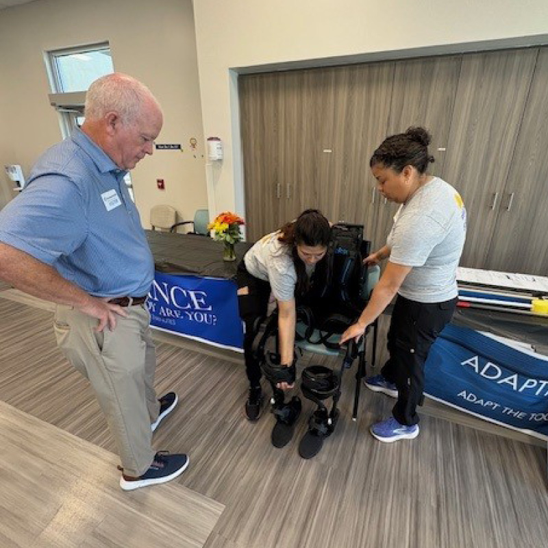 Two women demonstrate an exoskeleton device to a man at an indoor event, standing near a table with informational materials and a blue banner.
