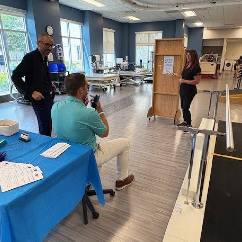 Three people in a rehabilitation therapy room, with one presenting beside a display board and therapy equipment visible in the background.