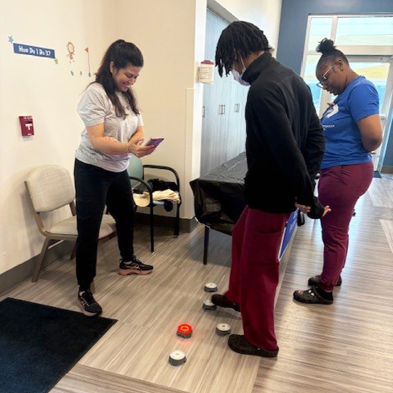 Three people participate in a therapy activity, stepping between floor markers in a clinical rehabilitation setting.