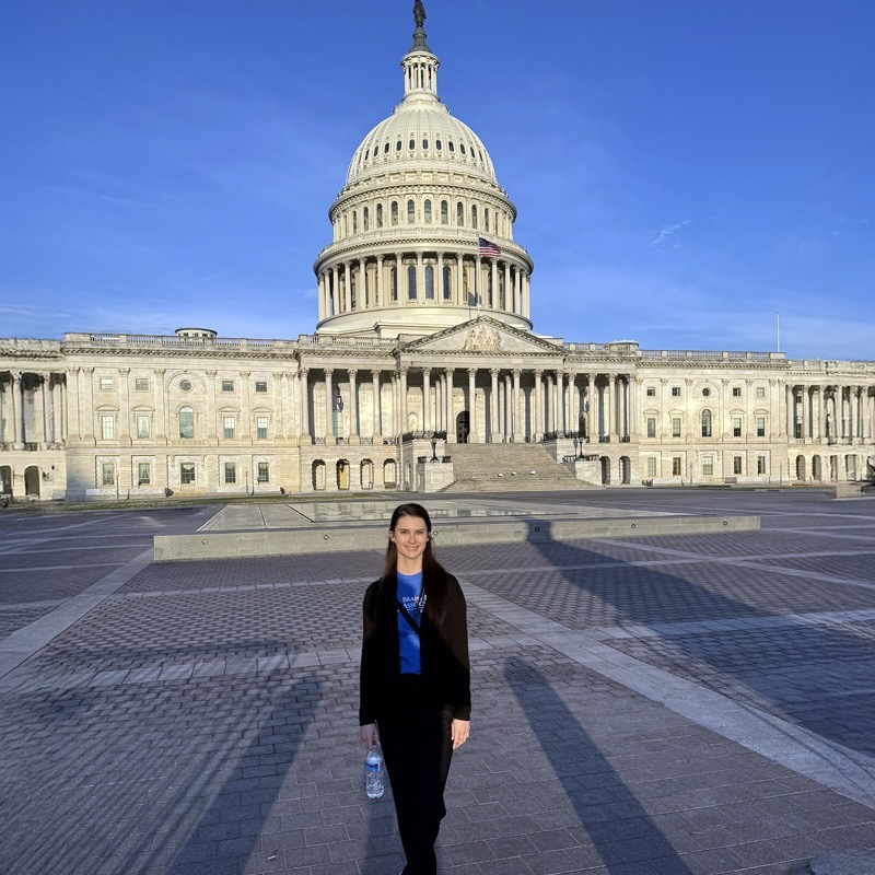 A woman stands holding a water bottle in front of the U.S. Capitol building under a clear blue sky. The scene conveys a sense of calm and openness.