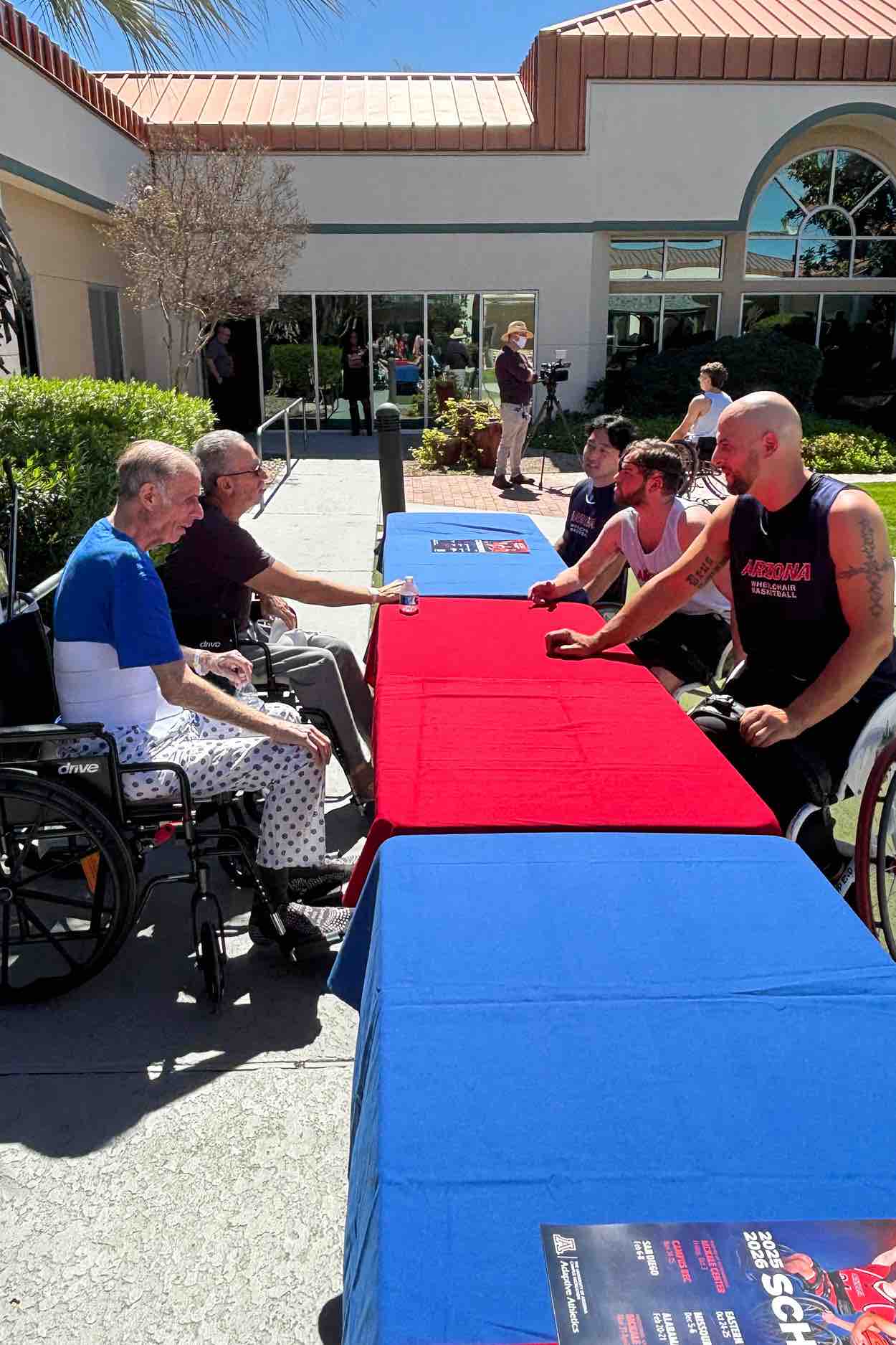Wheelchair basketball players and rehabilitation patients gather at an outdoor table, fostering connection, encouragement, and therapeutic engagement through adaptive sports within a hospital setting.