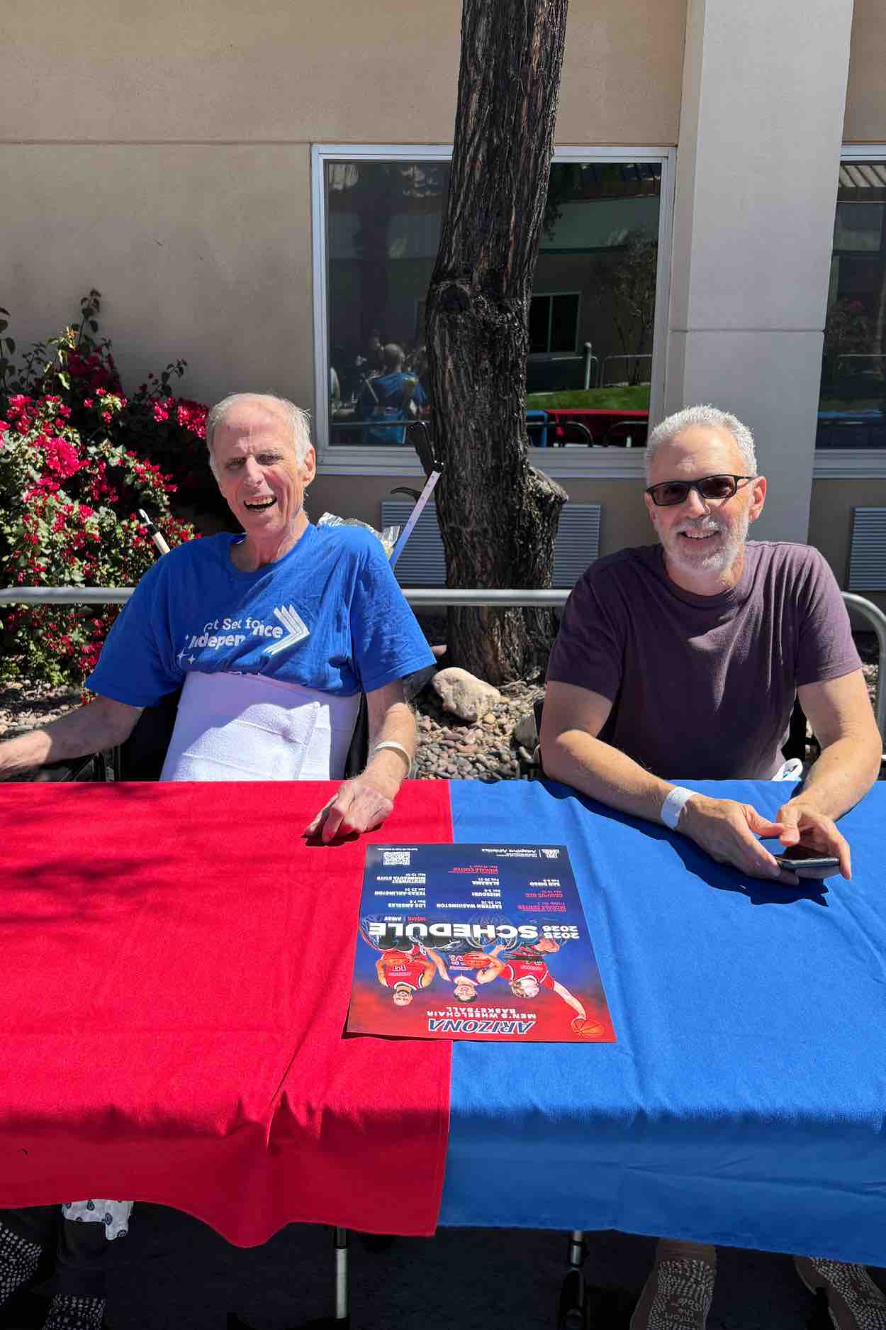 Two adults seated at an outdoor table smile during a wheelchair basketball event at a rehabilitation hospital, highlighting community engagement, adaptive sports support, and inclusive patient-centered care.