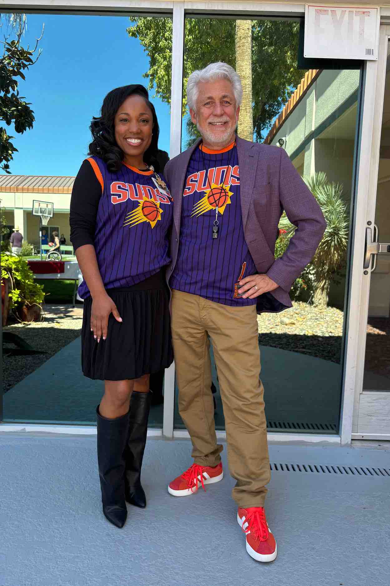 Two event supporters stand outdoors near a rehabilitation hospital wearing basketball jerseys, showing encouragement for wheelchair basketball programs that promote inclusion, motivation, and patient wellness.