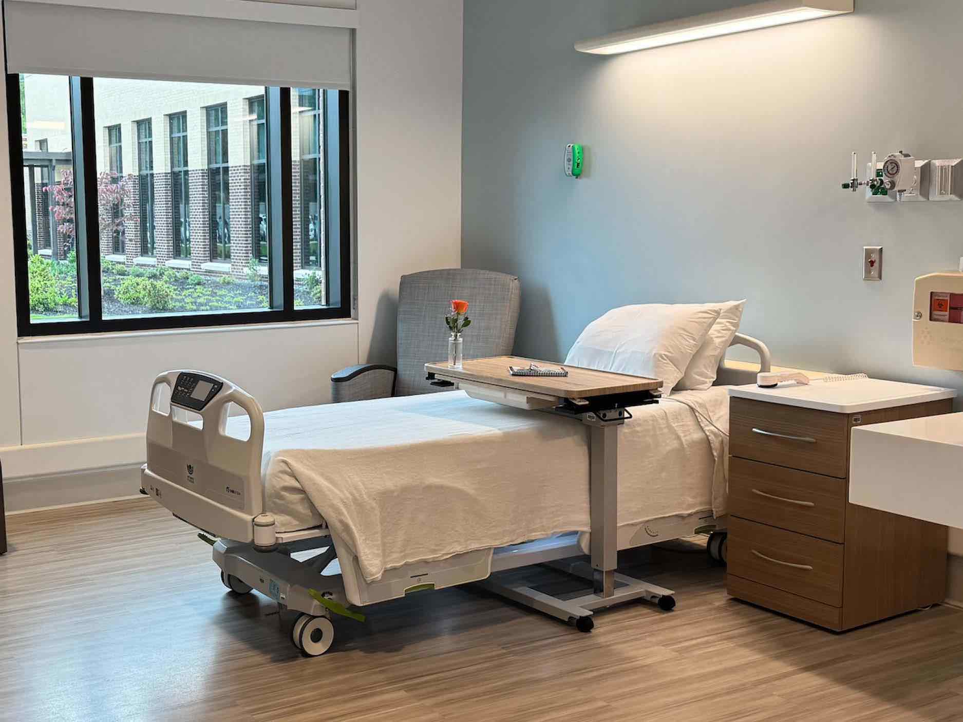 Private rehabilitation hospital patient room with adjustable bed, overbed table, recliner chair, window view and integrated medical equipment