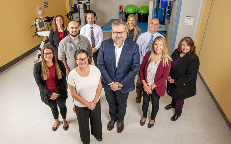 Encompass Health Erie’s professionals stand in a physical therapy clinic, with equipment like exercise balls and a treadmill. They are smiling, dressed in business and casual attire.
