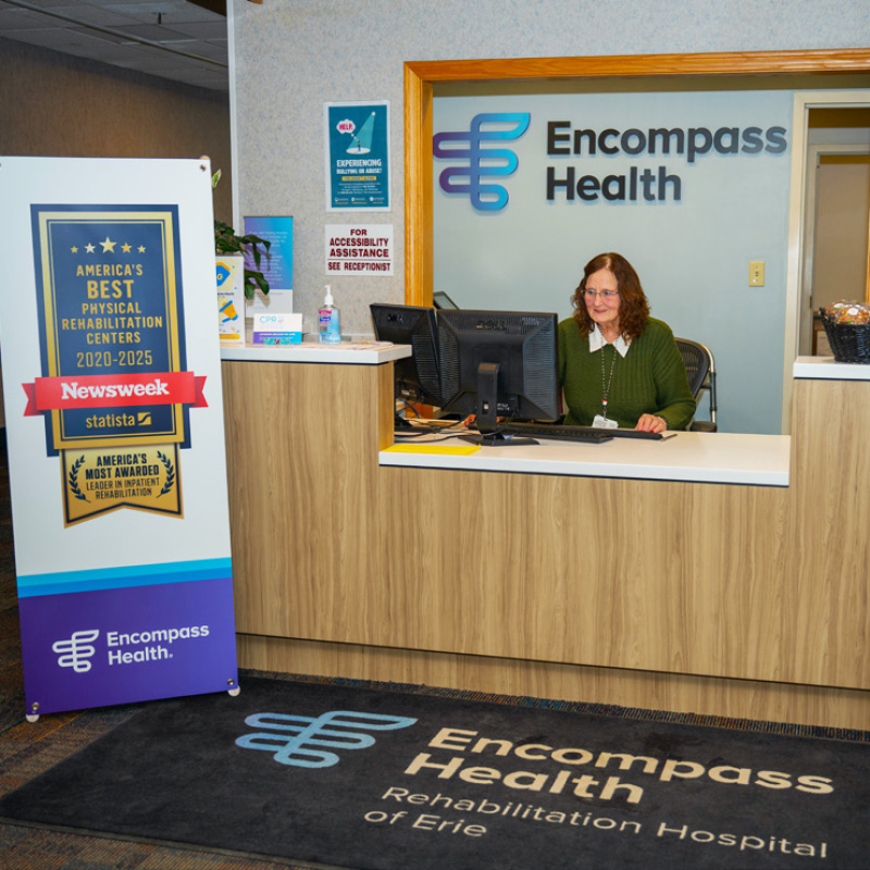 Reception area at Encompass Health with a smiling receptionist at a desk, large award sign to the left, and branded floor mat at the front.