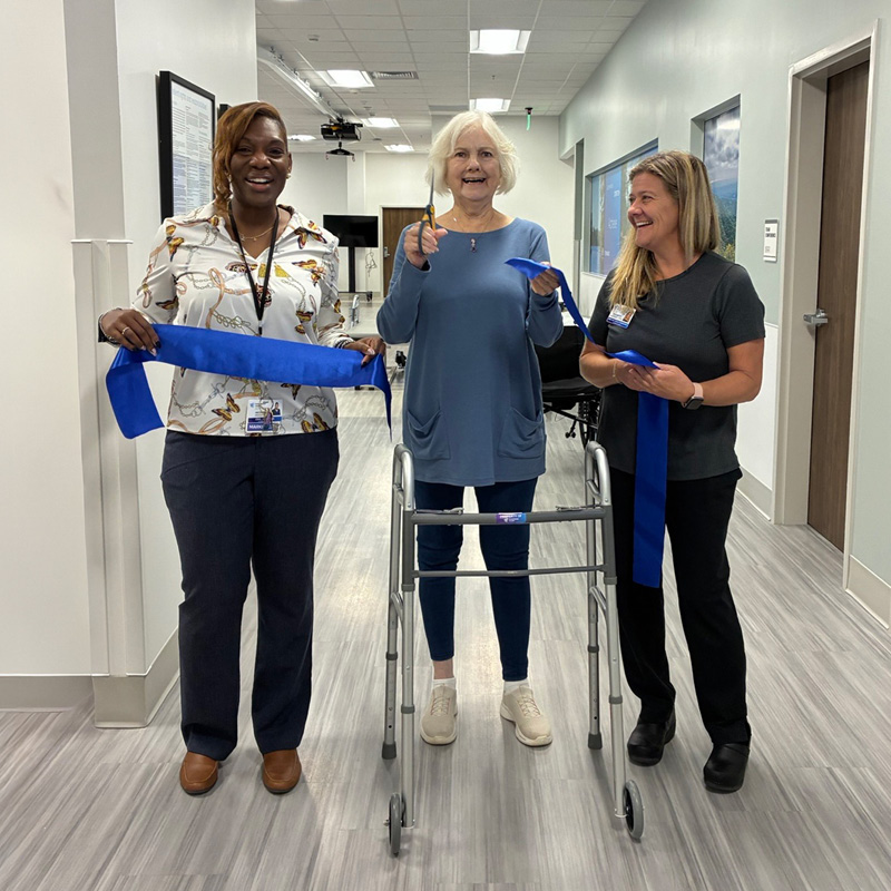 Three smiling women stand in a hallway holding a blue ribbon. The woman in the center, using a walker, cuts the ribbon with scissors, celebrating an event.