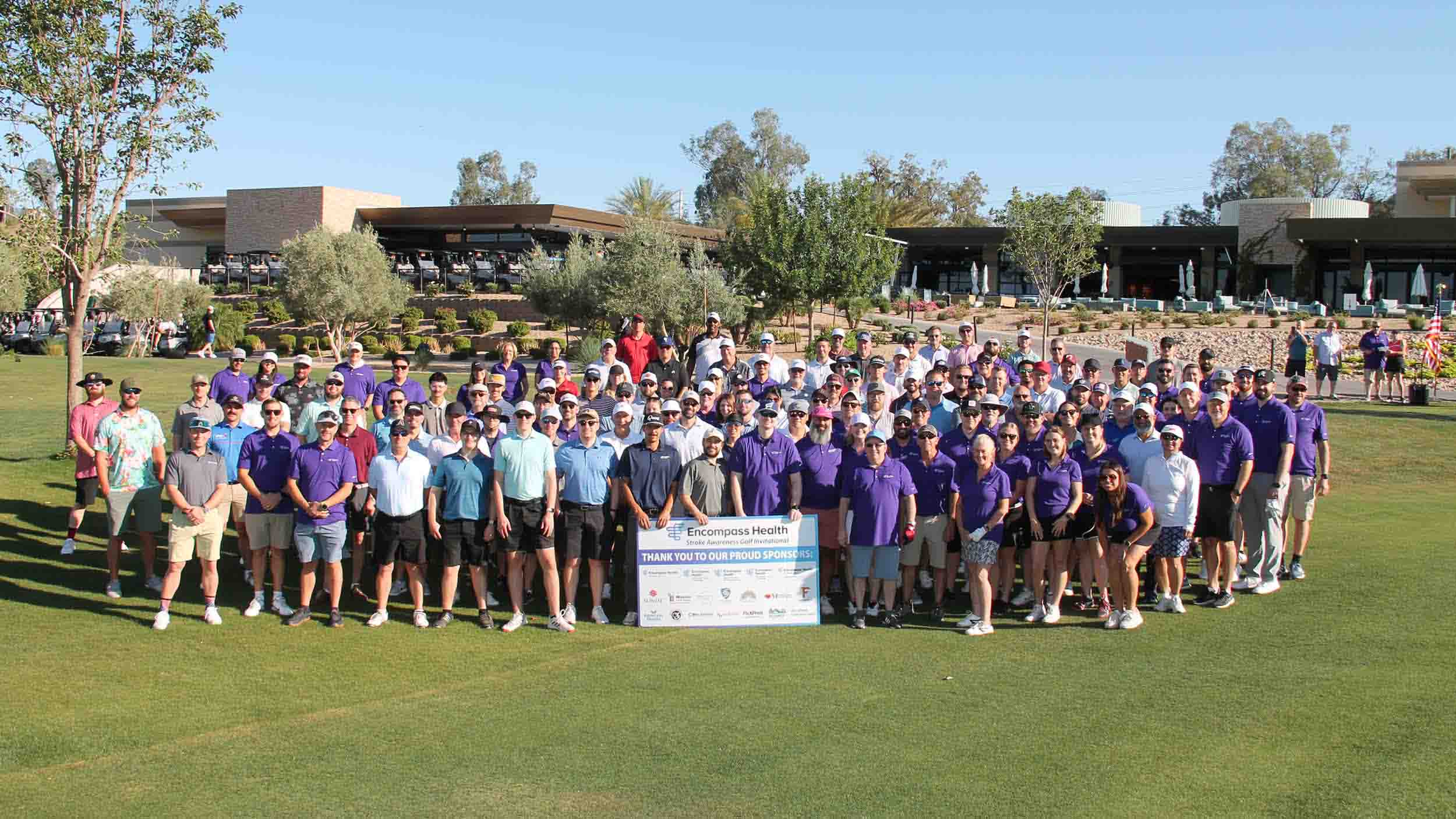 Large group of participants gather on a golf course for a veterans charity event, standing behind a sign recognizing sponsors, supporting rehabilitation programs and community outreach initiatives