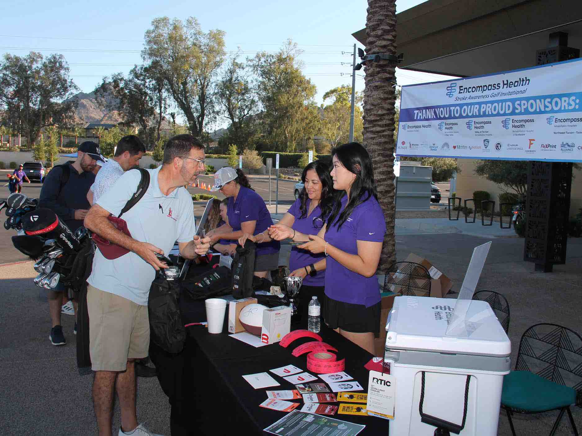 Event staff assist participants at a registration table during a veterans golf tournament, distributing materials and coordinating activities that benefit rehabilitation programs and patient care